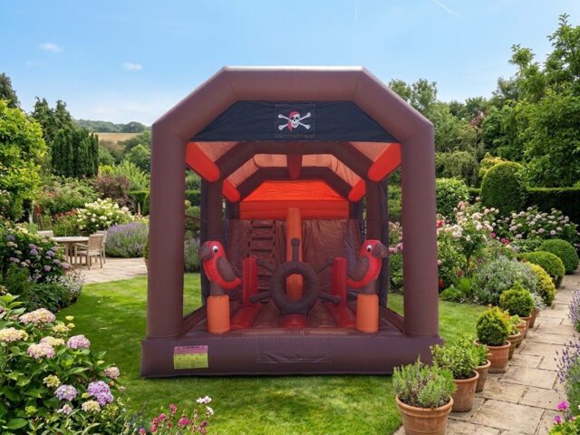 Combo hinchable de Piratas marrón y naranja con bandera pirata, loros, timón y tobogán en un jardín – Castillos Hinchables Madrid