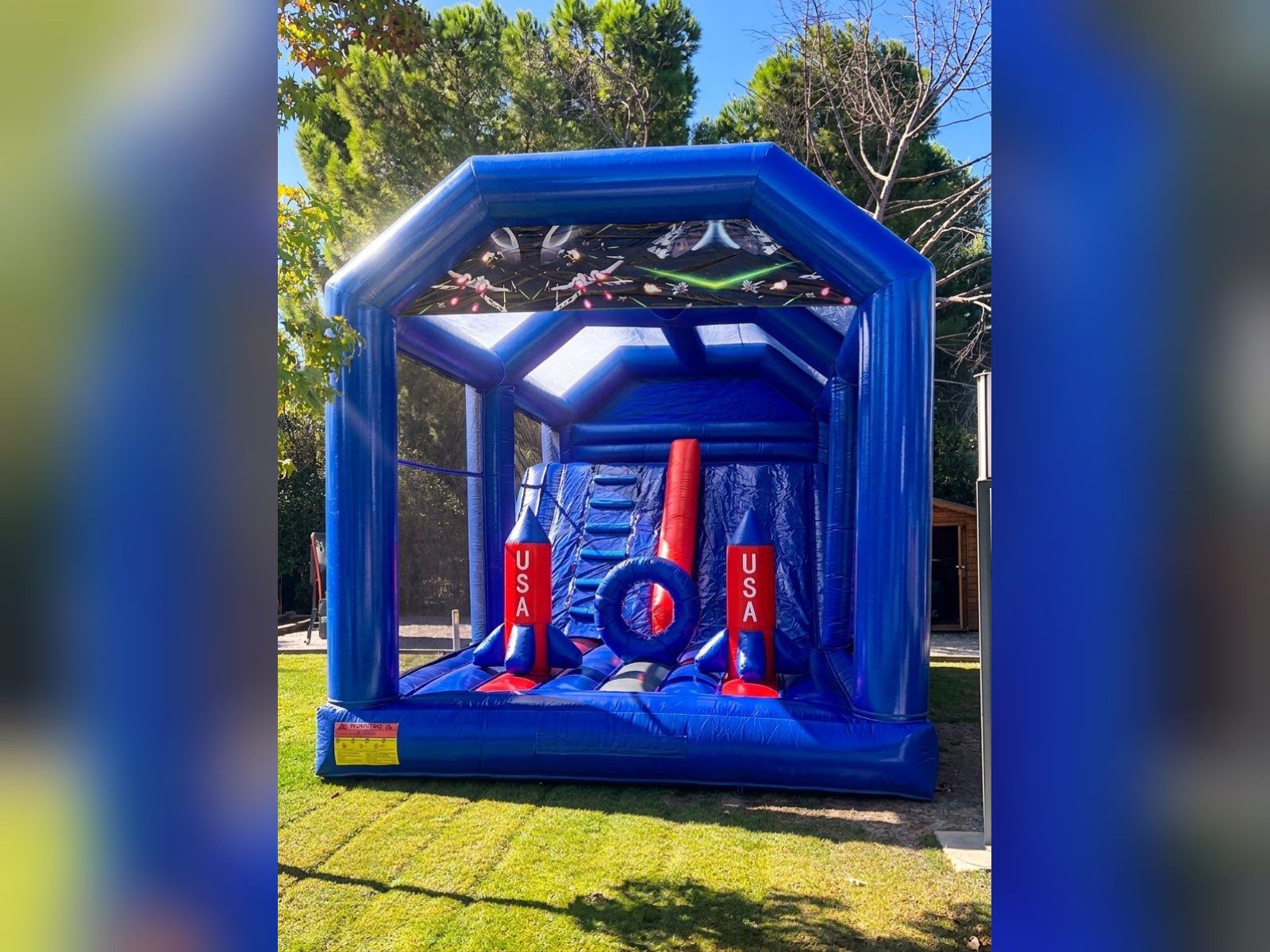 Combo hinchable Espacial XXL azul con cohetes rojos, techo con naves espaciales, obstáculos y tobogán en un jardín – Castillos Hinchables Madrid